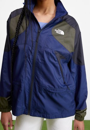 Henkilö, jolla on sinivihreä North Face -vetoketjutakki ja pitkät punotut hiukset, yhdistettynä valkoisiin housuihin.