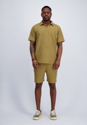 Man staat frontaal gekleed in een mosterdgele korte-mouwen shirt met bijpassende korte broek en witte sneakers tegen een effen achtergrond.