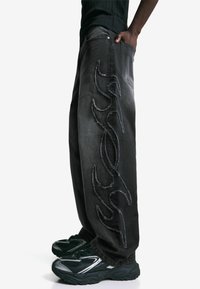 Vista laterale di jeans neri a gamba larga con ricami di fiamme alzati, indossati con sneakers nere e argentate e una camicia nera infilata dentro.