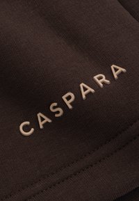 Brunt stoff med en teksturert overflate som har ordet "CASPARA" i lysebrune, pregete bokstaver. Sømdetaljer er synlige langs kanten.