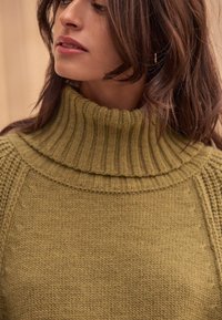 Olive green knitted sweater with a high ribbed collar, textured design, and wide cuffs. Features a layered neckline for added detail.