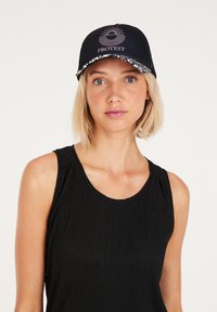 Cappello nero con un logo e un design solare in rosa, con un visiera decorata. Indossato da una persona con un taglio di capelli corto, su uno sfondo neutro.