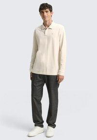 Beige Langarm-Polohemd mit Kragen, kombiniert mit dunkelgrauen Hosen und weißen Sneakers. Glattes Material, schlichtes Design, dezentes Logo-Detail.