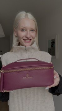 Trousse de maquillage bordeaux avec une finition texturée, fermeture éclair dorée et logo en relief. Design compact avec une poignée supérieure pour un transport facile.