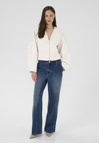 Hvid quiltet jakke med subtilt mønster, lynlås foran, kombineret med højttaljede blå denimjeans. Modellen har lyse sko på.