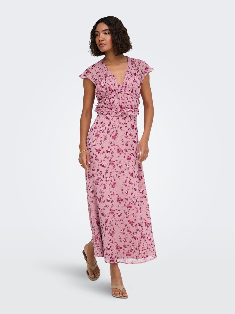 Femme portant une robe maxi fleurie rose avec des manches papillon, un décolleté en V et des sandales beiges, debout devant un fond blanc uni.