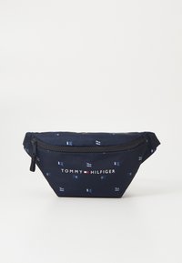 Laikku sininen kangaspussi valkoisella lippukuviolla, vetoketjukiinnitys ja "TOMMY HILFIGER" -logo edessä. Kompakti, kolmiomainen muoto.
