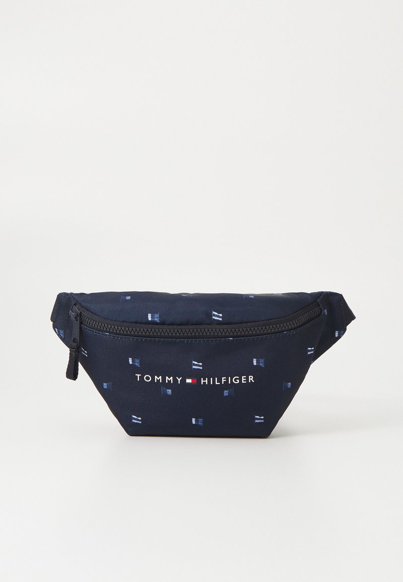 Laikku sininen kangaspussi valkoisella lippukuviolla, vetoketjukiinnitys ja "TOMMY HILFIGER" -logo edessä. Kompakti, kolmiomainen muoto.
