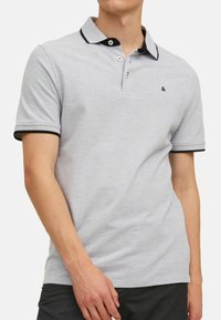 Lichtgrijs poloshirt met een twee-knoopsluiting, zwarte kraagafwerking en zwarte mouwen. Bevat een klein logo op de borst.