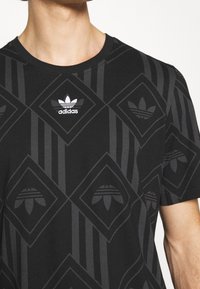 Camiseta negra de Adidas con patrones geométricos y múltiples logos Trefoil en gris oscuro y blanco en el pecho, llevada por una persona.
