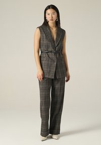 Gilet ajusté à carreaux marron et pantalon assorti, cintré à la taille, avec une subtile brillance. Talons aiguilles blancs et grandes boucles d'oreilles créoles.