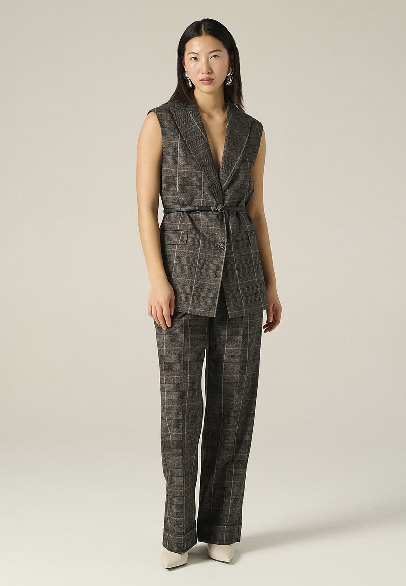 Gilet ajusté à carreaux marron et pantalon assorti, cintré à la taille, avec une subtile brillance. Talons aiguilles blancs et grandes boucles d'oreilles créoles.