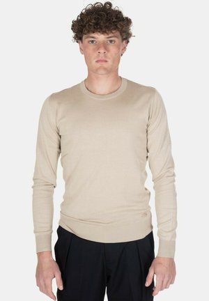 Giovane uomo con capelli ricci che indossa un maglione beige a maniche lunghe e pantaloni neri, in piedi davanti a uno sfondo chiaro e uniforme.