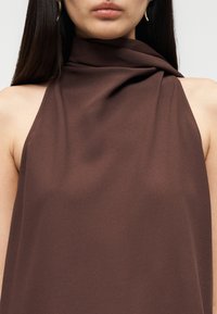 Καφέ τοπ halter από λείο ύφασμα, με ντραπέ λαιμόκοψη και χωρίς μανίκια. Αναδεικνύει τους ώμους με καθαρή, μινιμαλιστική γραμμή.