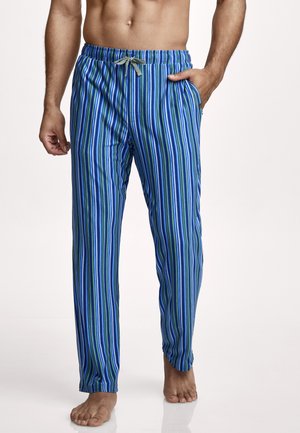 Mann trägt blau-grün-weiß vertikal gestreifte Pyjamahose mit grauem Kordelzug, steht barfuß mit einer Hand in der Tasche vor weißem Hintergrund.