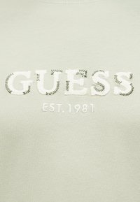 Λευκά γράμματα "GUESS" με πράσινη ραφή και κεντημένο "EST. 1981" από κάτω σε ανοιχτοπράσινο ύφασμα.