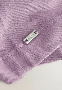 Close-up of lavender knit fabric with a small rectangular metal tag engraved with "NEXT" sewn near stitched seam.