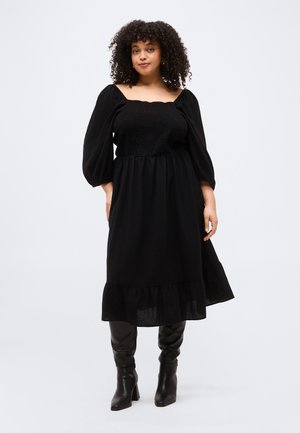 Femme aux cheveux bouclés portant une robe noire smockée à épaules dénudées avec des manches bouffantes et des bottes à talons noires, debout devant un fond uni.