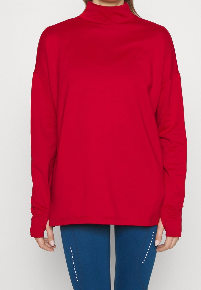 Femme portant un haut ample rouge à col roulé et manches longues, associé à des leggings bleus avec des points réfléchissants verticaux sur les cuisses.