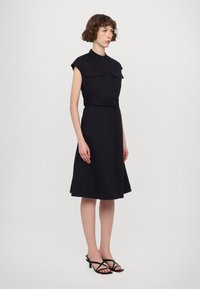 Crna haljina bez rukava s prednjim kopčanjem na dugmad, džepovima s preklopom, strukom s remenom i A-krojem, uparena s crnim sandalama s trakama.