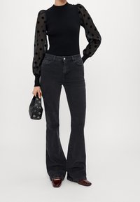Donna che indossa un top nero aderente con maniche a forma di palloncino trasparenti a pois, jeans scuri a zampa d'elefante, mentre tiene in mano una piccola borsa nera con occhielli metallici.