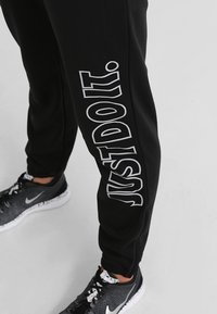 Zwarte joggings met een gladde stof hebben een grote witte "JUST DO IT." opdruk langs de zijkant, met elastische boorden en een comfortabele pasvorm.