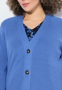 Modrý pletený cardigan s výstřihem do V a ribkovanou texturou; má dva tmavé knoflíky vpředu. Nosit přes černé tričko s modrým nápisem.