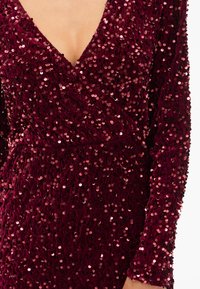 Burgundská večerná šaty so zdobením, s dlhými rukávmi a hlbokým výstrihom do V, vyrobené z textúrovaného materiálu, ktorý je posiaty trblietavými okrúhlymi flitrami.