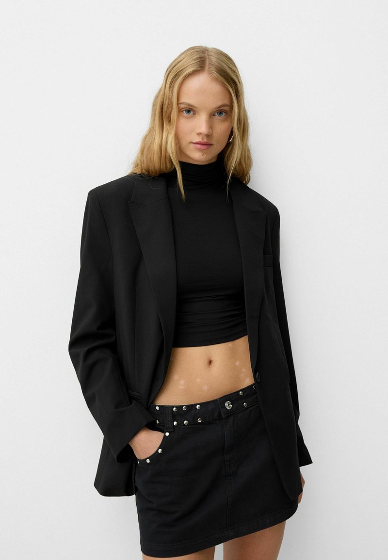 Veste oversize noire sur un col roulé noir et une mini-jupe noire ajustée, avec des clous argentés sur la taille et une texture lisse.