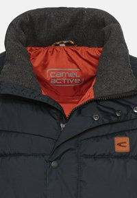 Marineblaue gepolsterte Jacke mit einem grauen Fleecekragen und orangefarbener Innenausstattung. Sie verfügt über silberne Reißverschlüsse, Druckknöpfe und ein gefertigtes Lederlogo auf der Brust.