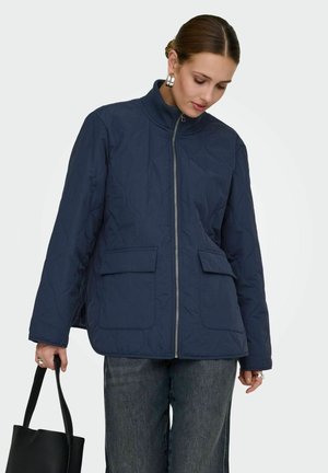 Mujer con chaqueta acolchada azul marino y vaqueros oscuros, sosteniendo un bolso tote de cuero negro, mirando hacia abajo contra un fondo liso.