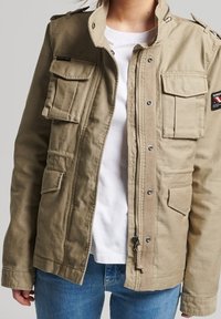 Beige militærinspireret jakke med flere lommer foran, trykknapper, lynlås og skulderstropper. Båret over en hvid skjorte.