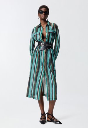 Femme portant une robe chemise rayée bleu canard, noir et marron avec une large ceinture noire, des ballerines noires à lanières, de grandes lunettes de soleil et des boucles d’oreilles créoles.