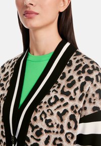 Kardigan mit Leopardenmuster und schwarz-weiß gestreiftem Kragen, mit Knopfverschluss. Über einem leuchtend grünen Oberteil getragen. Weicher Stoff.