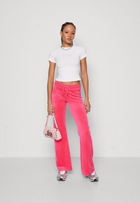 Rosa sammetsbyxor med utsvängd nederdel och dragsko i midjan, tillsammans med en figurnära vit t-shirt med pr präglat varumärke. Liten rosa handväska ingår.