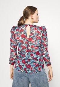 Blusa floreale con maniche a sbuffo, caratterizzata da un motivo di fiori blu e rossi su uno sfondo trasparente, e una chiusura a oblò sul retro.
