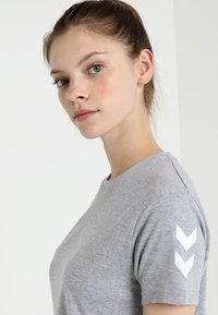 Junge Frau mit heller Haut und zurückgebundenem Haar, die ein graues T-Shirt mit zwei weißen Chevron-Pfeilen am Ärmel trägt und seitlich schaut.