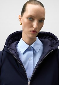 Granatowa kurtka z kapturem i pikowanym wewnętrznym podszewką, zestawiona z jasnoniebieską koszulą zapinaną na guziki. Posiada zamek z przodu oraz złote kolczyki w kształcie kółek.