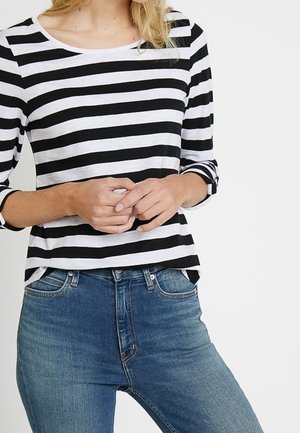 Femme portant un haut à manches longues à rayures horizontales noires et blanches et un jean bleu, les mains jointes lâchement à la taille.