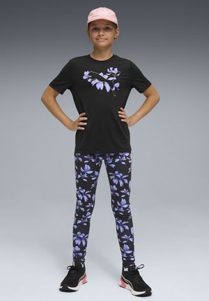 Młoda osoba stojąca z rękami na biodrach, ubrana w różową czapkę Puma, czarną kwiatową koszulkę Puma, dopasowane legginsy oraz czarne tenisówki.