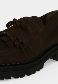 Mocassin en suède brun foncé avec semelle épaisse en caoutchouc noir et détail à lacets effilochés sur le dessus.