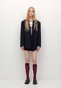 Schwarzer Nadelstreifen-Oversized-Blazer über einem weißen Hemd mit pinkem Design, kombiniert mit einem schwarzen Minirock, bordeauxroten Kniestrümpfen und schwarzen Schuhen.