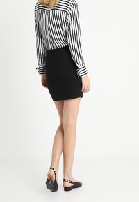 Donna che indossa una minigonna nera, scarpe slingback nere con borchie e una camicia a righe verticali bianco e nero a maniche lunghe, in piedi su uno sfondo bianco.