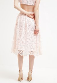Falda midi de encaje en rosa claro con un patrón floral, que presenta un dobladillo en forma de ola. Combinada con zapatos de tacón nude.