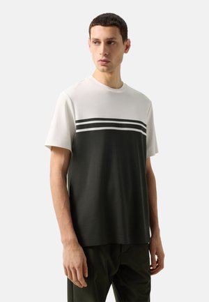 Mann trägt kurzärmeliges, schwarz-weißes Shirt mit zwei schwarzen Streifen über der Brust, steht vor einfarbigem Hintergrund.