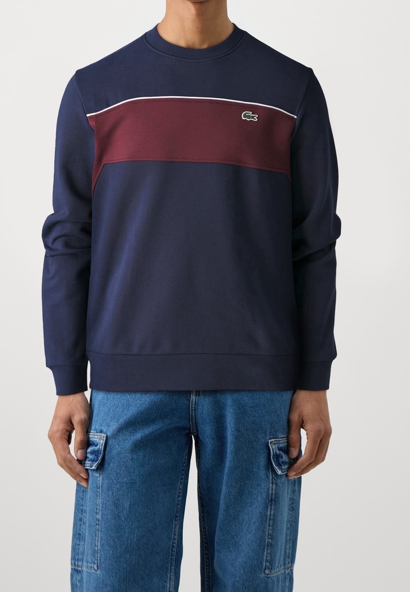 Felpa navy con striscia orizzontale bordeaux, piping bianco e piccolo logo verde. Presenta collo a giro e polsini a coste; texture liscia.