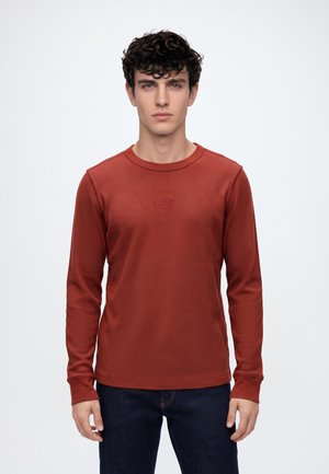 Jeune homme portant une chemise à manches longues couleur rouille avec le logo "GAP", debout devant un fond clair uni.