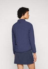 Navy blue fabric jacket with a collar, long sleeves, and side pockets. Features a textured surface and a small logo at the back neckline.