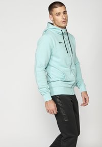 Sudadera con cremallera de color azul claro, con capucha ajustable, bolsillos frontales y puños de canalé; combinada con pantalones de chándal negros que presentan un logo.