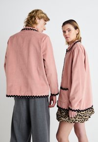 Ružové kordové bundy s čiernym ozdobným lemovaním, ktoré majú voľný strih. Mužský model ich kombinuje so šedými nohavicami a ženský model nosí sukňu s leopardím vzorom.